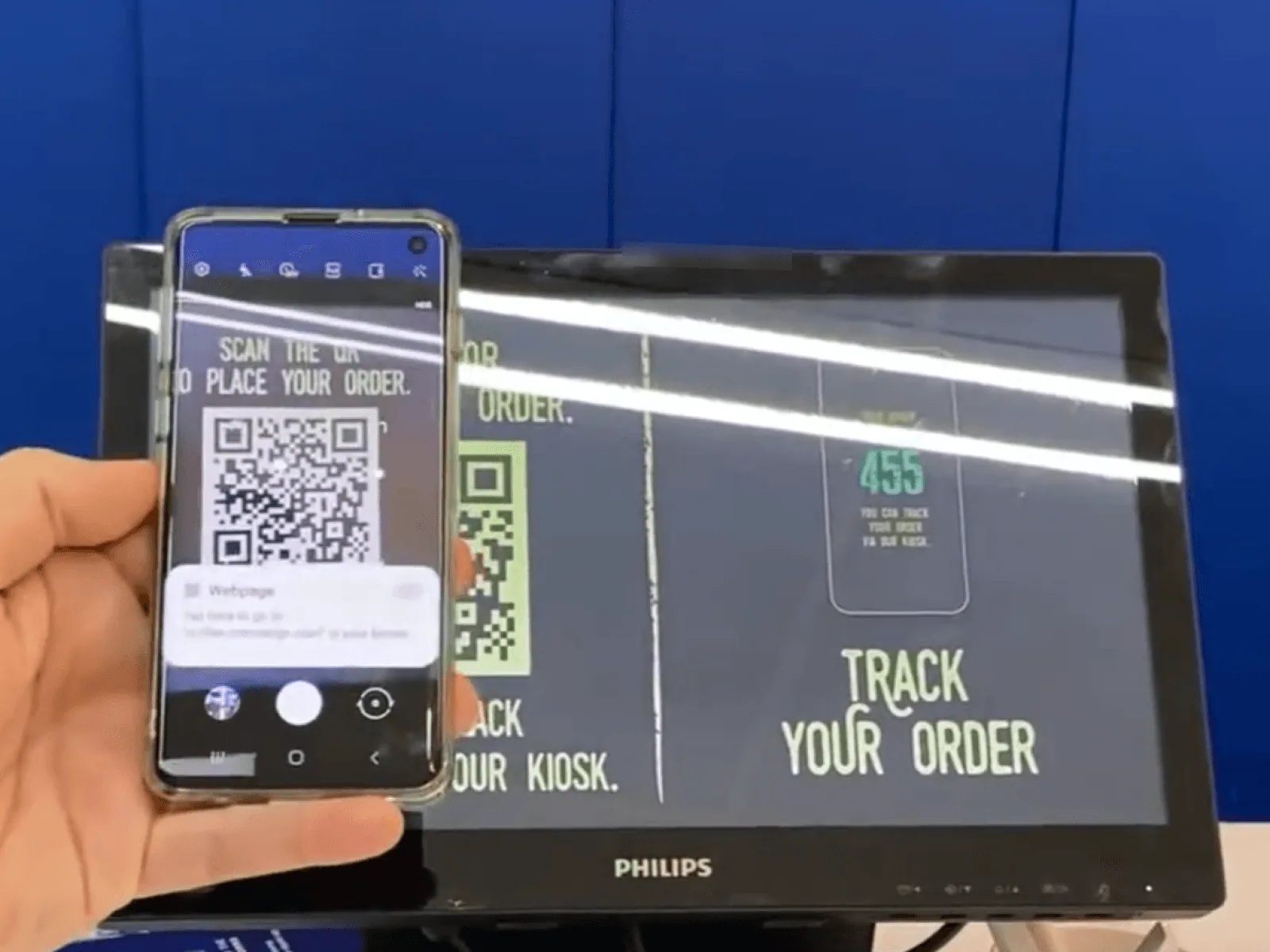 A guest scanning the screen's QR code to place an order from their phone, beside the live 'Track your order' display