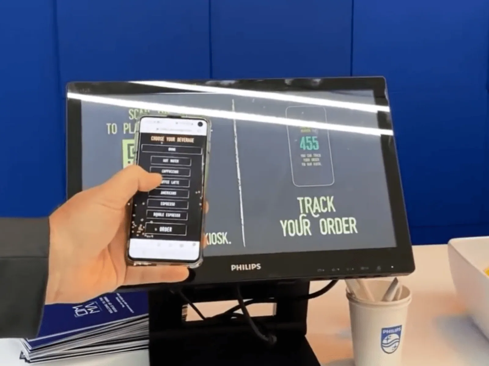 A guest choosing items on their phone while the order-tracking screen and a finished coffee sit on the counter